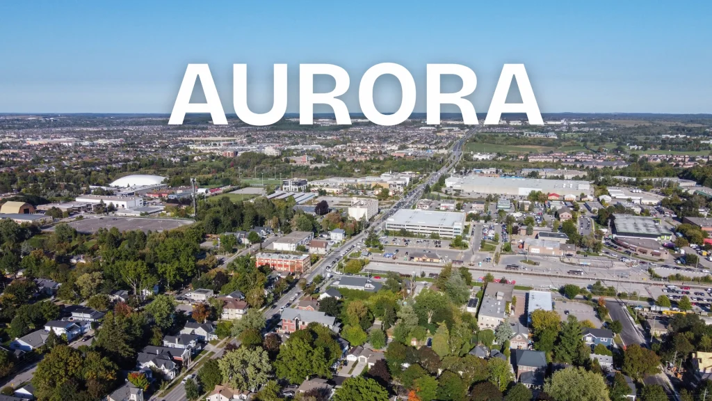 High-angle aerial view of Aurora, Ontario, showing residential neighborhoods and commercial areas under a clear blue sky with "AURORA" text overlay.