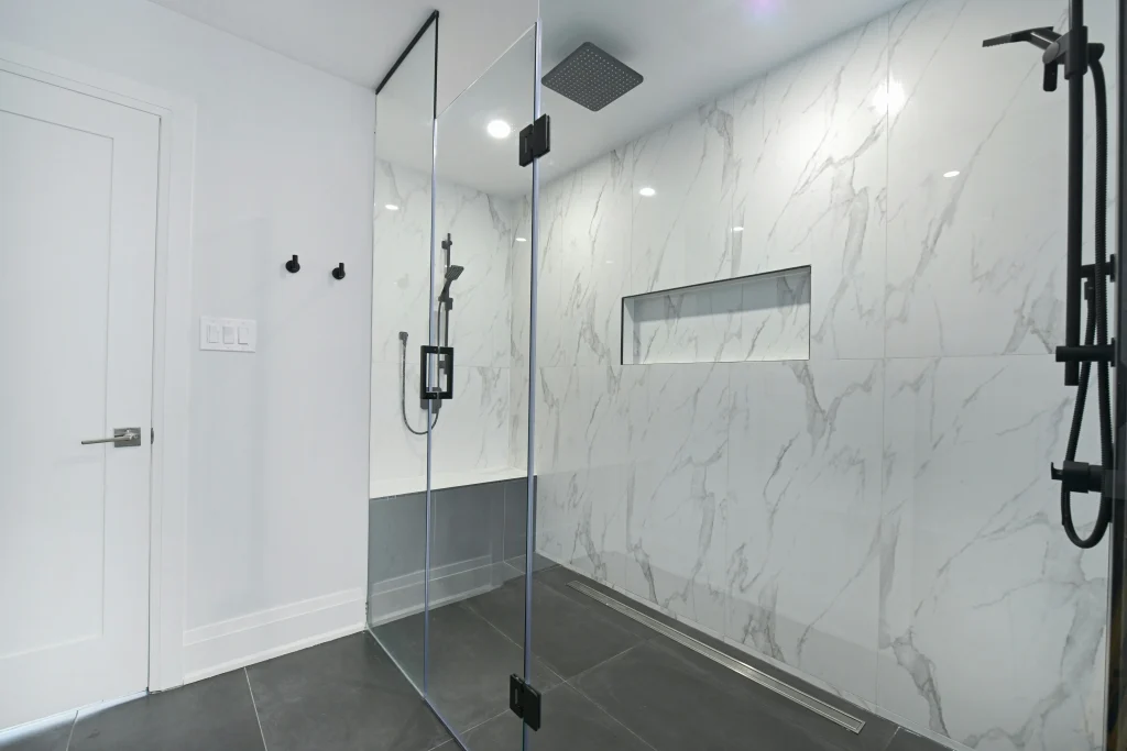 Modern bathroom renovation in Aurora featuring a walk-in glass shower with marble-patterned wall tiles, a recessed storage niche, matte black rainfall showerhead, and dark grey floor tiles.