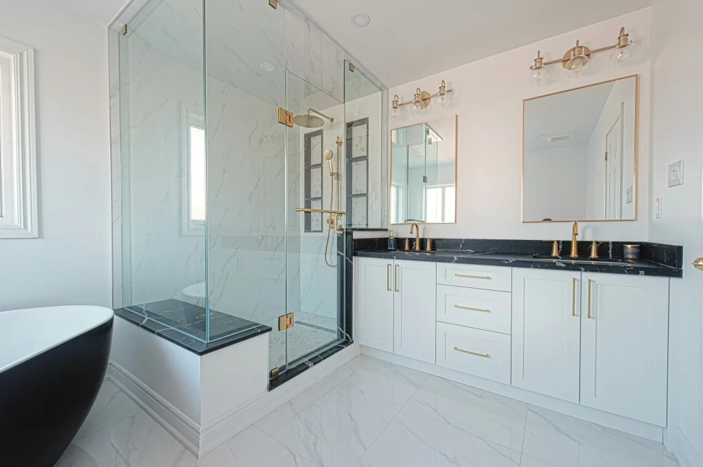Luxury master bathroom renovation in Aurora featuring a double white vanity with black marble countertops, gold hardware, a glass-enclosed walk-in shower with a marble bench, and a black soaking tub.