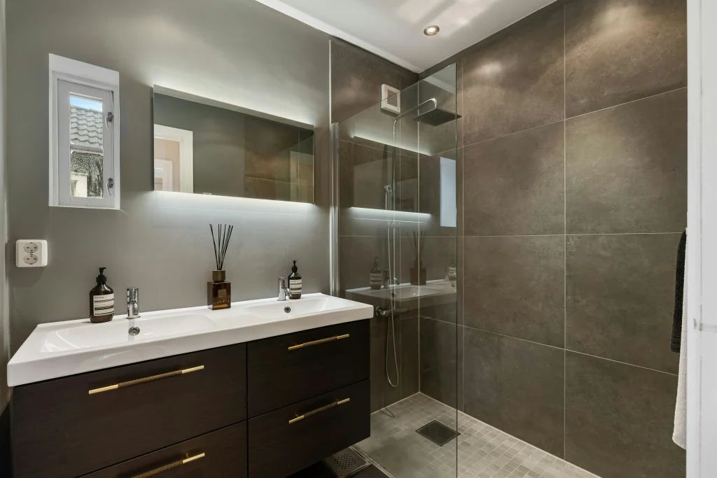 Modern bathroom renovation in Aurora featuring a dark wood double vanity with gold handles, a backlit mirror, and a walk-in glass shower with large-format charcoal tiles and mosaic flooring.