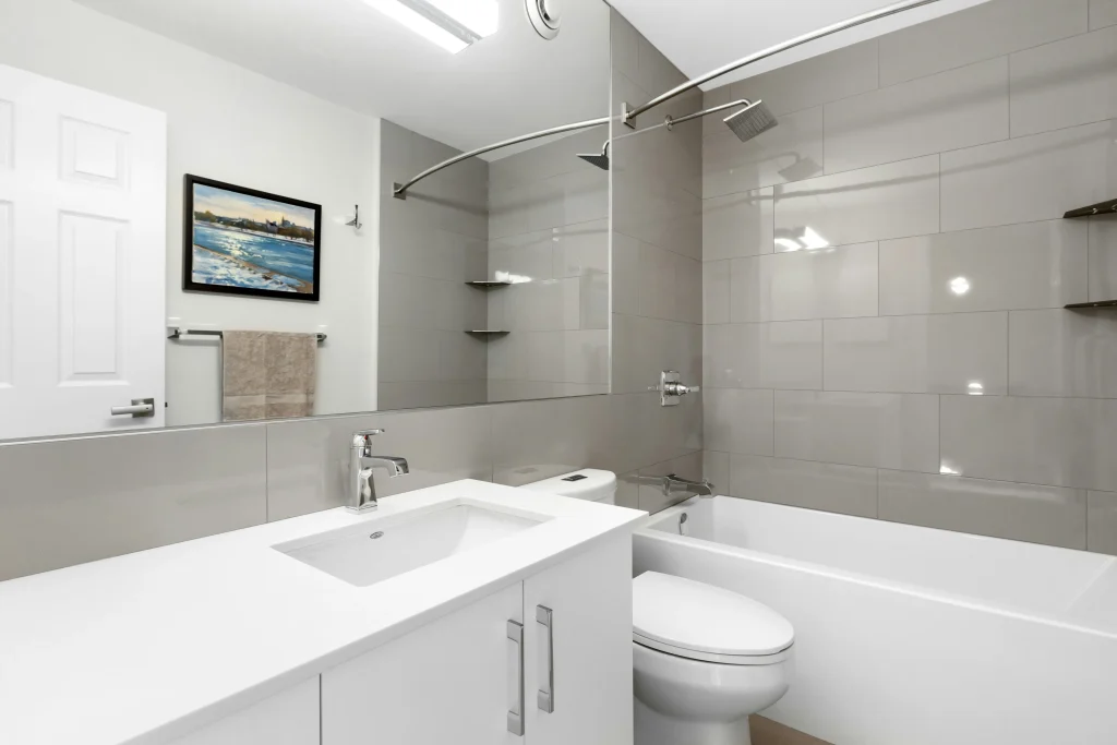 Bright basement bathroom renovation in Aurora, Ontario, featuring a white single-sink vanity, a large wall mirror, and an alcove bathtub with glossy grey subway-style wall tiles.